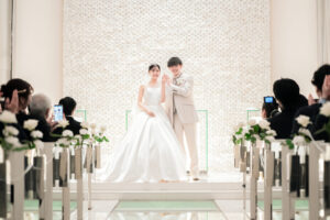 白を基調としたチャペルで結婚指輪を披露する新郎新婦。ゲストの拍手に包まれながら、祭壇の前で笑顔を見せるドレスとタキシード姿の挙式シーン。