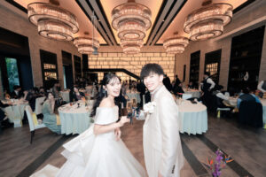 豪華なシャンデリアが輝く披露宴会場でグラスを持ち笑顔で乾杯する新郎新婦。ゲストが集まるウェディングパーティーで幸せいっぱいの表情を見せる二人。