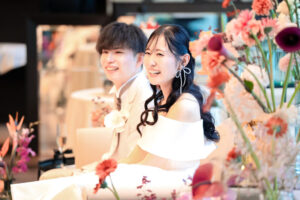 披露宴の高砂席で笑顔を見せる新郎新婦のウェディングフォト。手前に飾られたカラフルなメイン装花が華やかな、アットホームな結婚式パーティーの風景。
